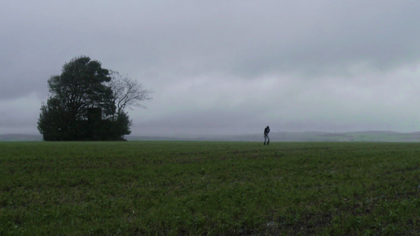 Une personne traverse un champ sous la pluie près d’un grand arbre, dans Le Corps des Anges.