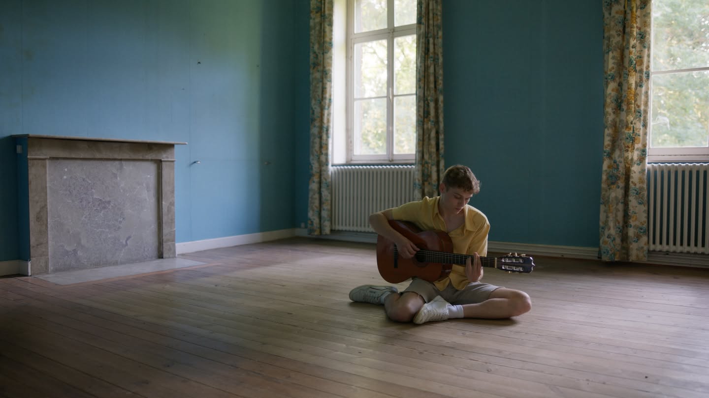 Charlie joue de la guitare, assis dans une grande pièce bleue.